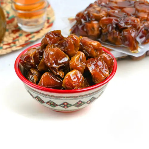 Rote und weisse Schale mit Premium gepressten Sukari-Datteln, dahinter eine volle Packung, im Hintergrund weitere Datteln und oben links ein Glas mit arabischem Kaffee auf einer bunten Matte.