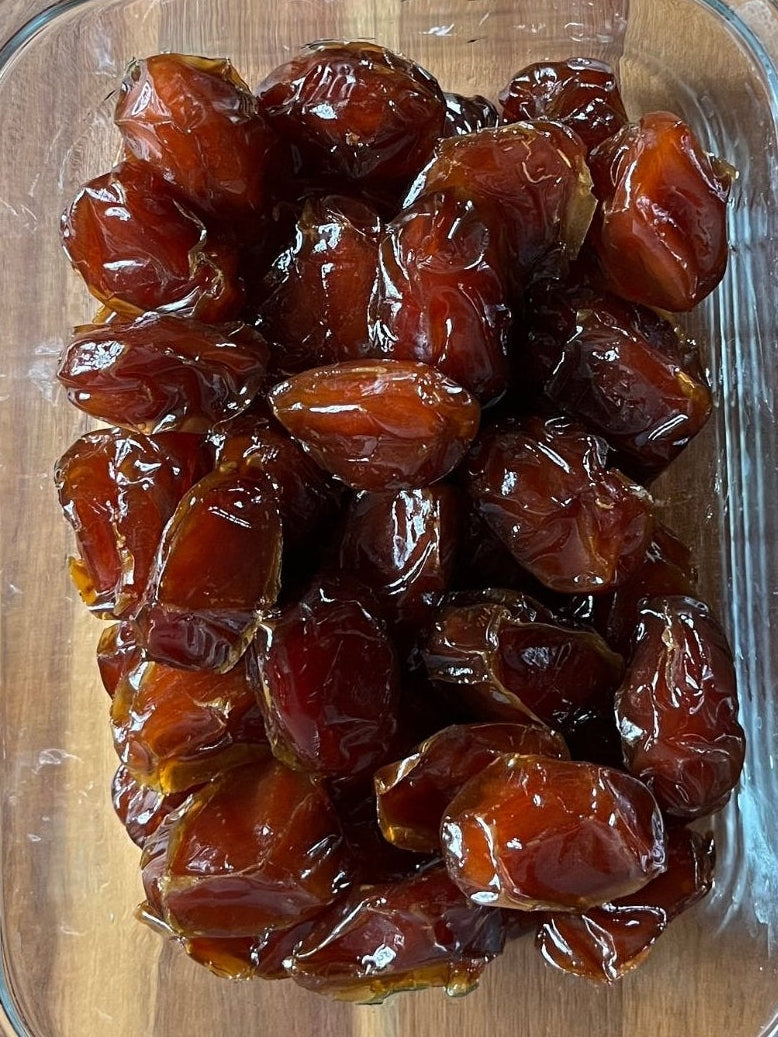 Glass container of dates on a wooden surface with a bowl of nuts and fruit in the background.